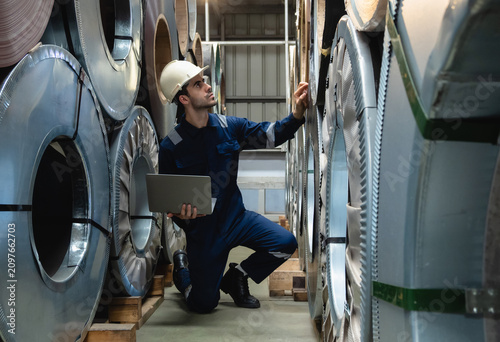 professional male industrial inspector in navy blue uniform and hardhat kneels with laptop, conducting an inventory check on large steel coils inside modern manufacturing factory warehouse.