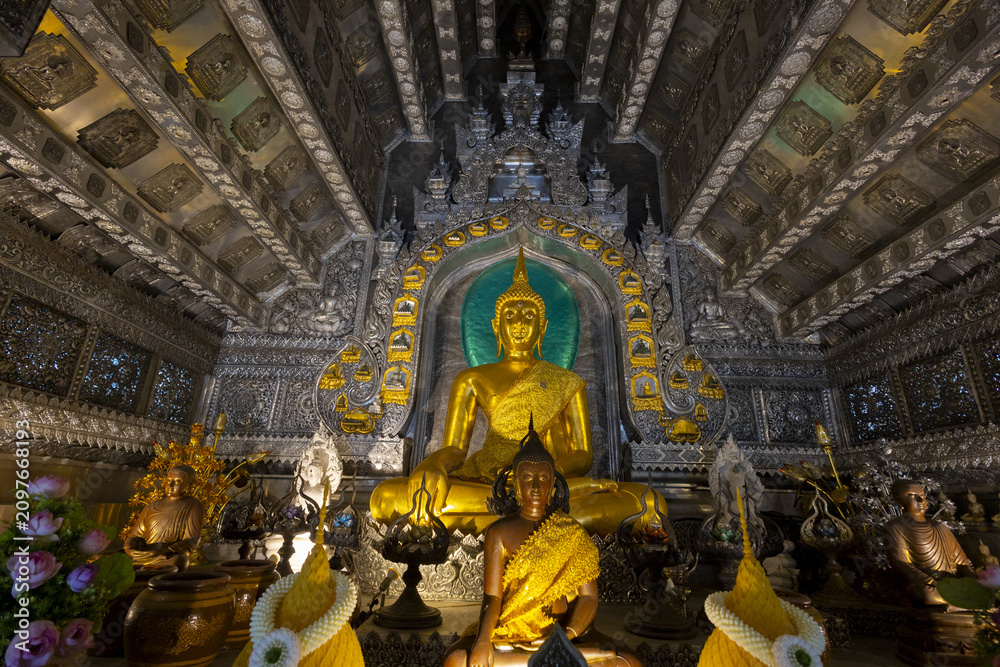 Obraz premium Golden Buddha statue seated in ornate silver temple interior with intricate carvings, lotus offerings and ceremonial decorations conveying serene sacred atmosphere and cultural reverence