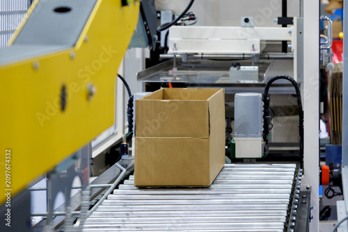 brown cardboard box on roller of conveyor belt at automatic carton sealing machine. Industrial product packaging, logistics, and e-commerce fulfillment factory automation concept
