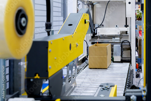 brown cardboard box on roller of conveyor belt at automatic carton sealing machine. Industrial product packaging, logistics, and e-commerce fulfillment factory automation concept