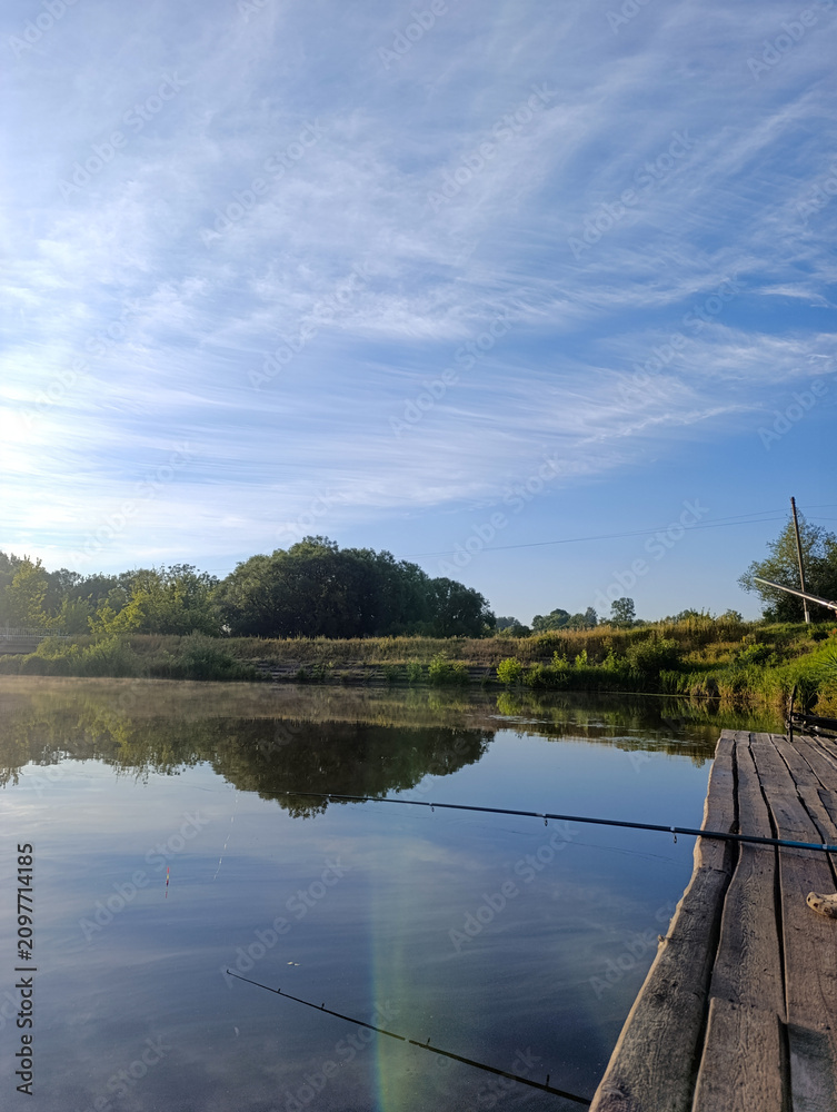 Fototapeta premium Fishing rods on a wooden pier by a calm river on a sunny morning