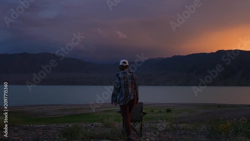A back view of a woman walking to a camping chair and sitting down to watch a mountain lake landscape at twilight. High-quality B-roll footage for travel advertising, digital content creation, and lif