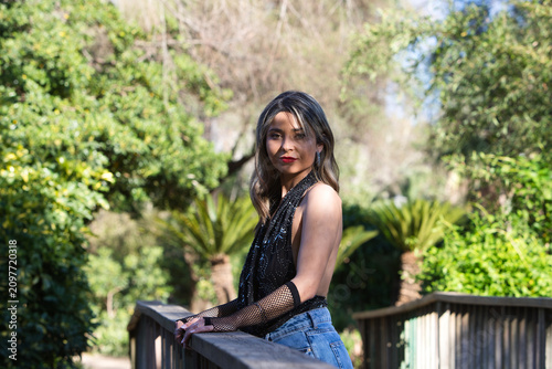 A young, attractive woman, wearing make-up and dressed in jeans, gloves and a black T-shirt, leaning on the railing of a wooden bridge surrounded by lush greenery.