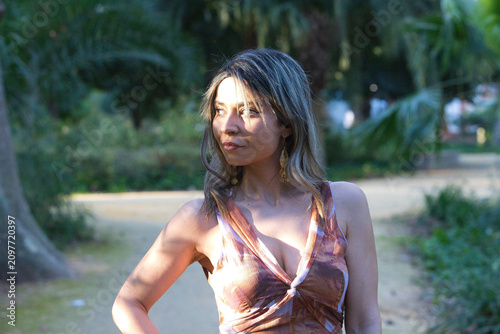 A young, pretty woman in a floral-print dress walks through the park amongst the trees, striking various poses and striking a pose like a model, whilst enjoying the natural surroundings.