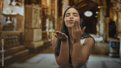 Foto Young hispanic woman blows a kiss with raised hand to lips in an ornate building hall, puckered lips and casual denim visible; affection warmth
