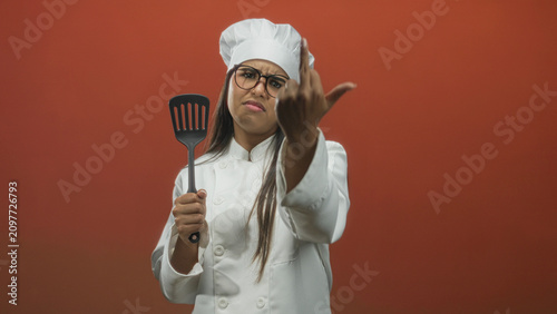 Obraz na płótnie Woman chef in white coat holds spatula and extends middle finger toward camera in a studio with red backdrop, face frowning; defiance attitude