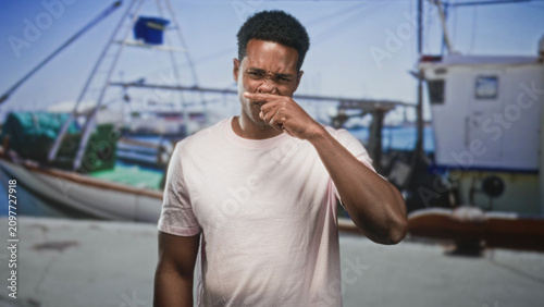 Photos Man touching mouth with finger beside fishing boat on dock, hand near lips and pained expression; disgust unease
