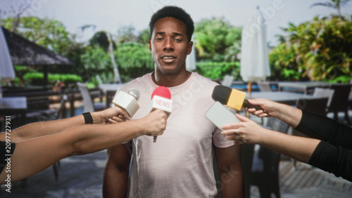 Bild auf Leinwand Man speaking into multiple microphones and a smartphone held by reporters on a restaurant patio outside a building; press scrutiny calm