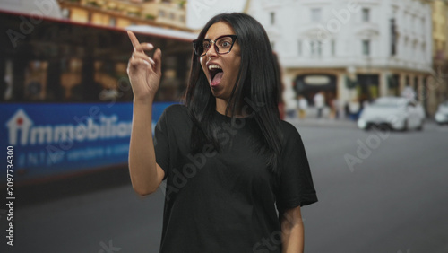 Fotografía Woman with crossed arms smiling, wearing glasses, on a busy city street beside a red bus and classic buildings; confidence independence