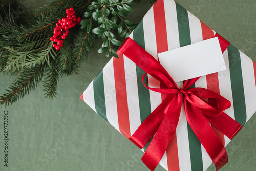 High-angle close-up of a striped Christmas present with a red ribbon bow and an empty gift tag mockup next to evergreen branches with red berries.