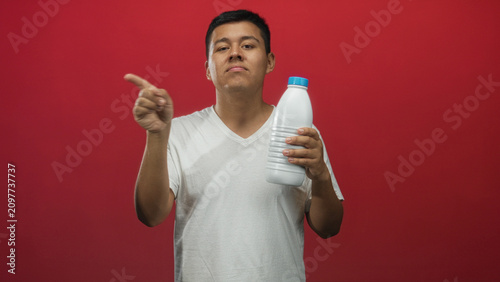 Foto Young hispanic man holding white plastic bottle with blue cap points finger toward camera in red studio; confidence endorsement