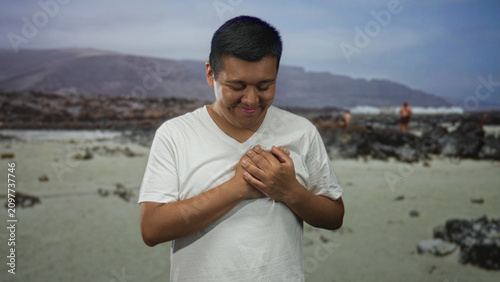 Fotografía Man holding hands to chest on a sandy beach with closed eyes and a soft smile, rocky shoreline and swimmers in the distance; calm gratitude