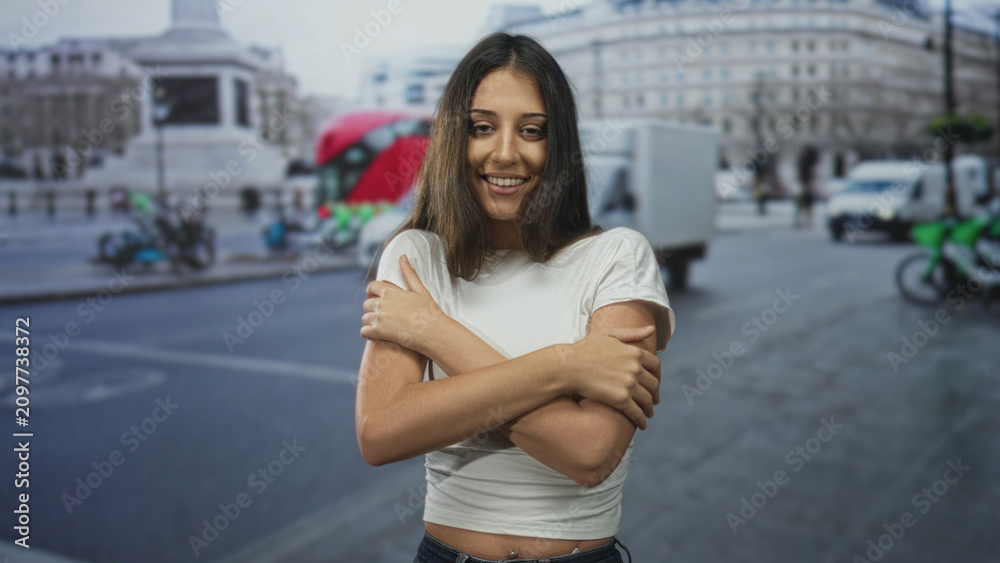 Fototapeta premium Young hispanic woman hugging bare shoulders with arms crossed on busy city street among passing vans and buses; self care contentment.