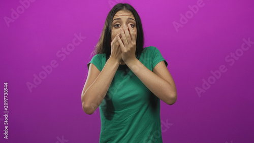 Bilde på lerret Young woman in green shirt with hands covering mouth, wide eyes and visible gasp in purple studio; surprise concern