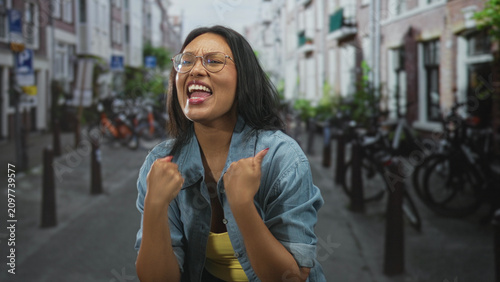 Принти на полотні Woman with clenched fists and open mouth cheering on a street lined with bicycles and rowhouses; triumph elation