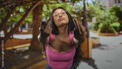 Konstfotografi Young hispanic woman in pink tank top shows middle fingers toward the camera on a street plaza; defiance attitude