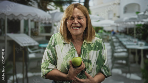 Canvastavla Woman holding green apple in her hand smiling and laughing at an outdoor cafe by a white building with umbrellas and tables; joyful warmth