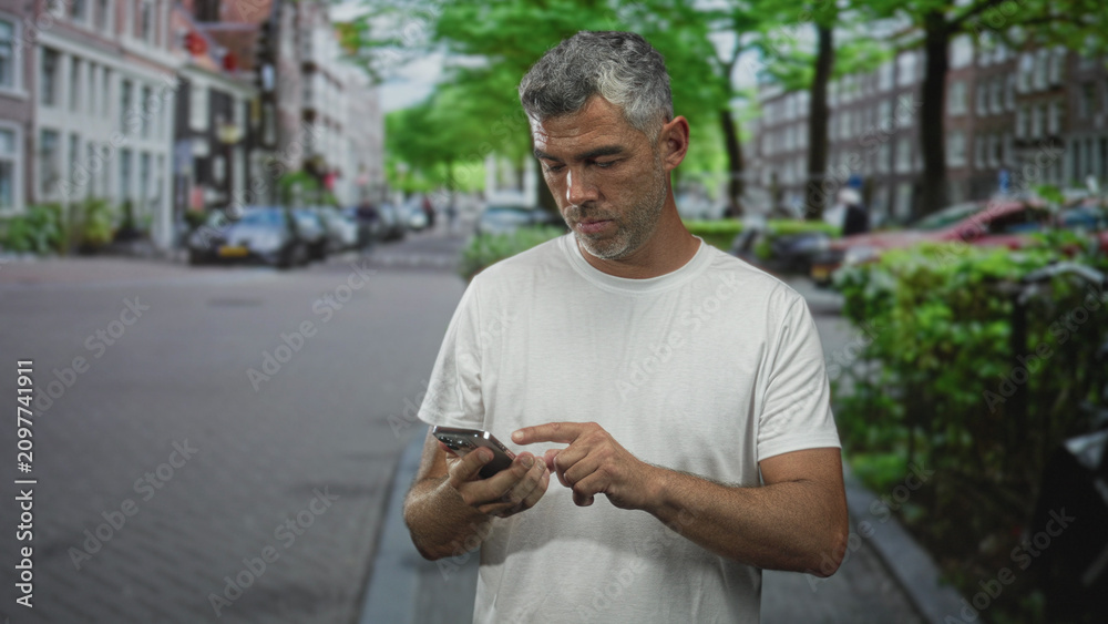 Fototapeta premium Middle age man with grey hair holds smartphone to ear on a cobbled city street, forearm exposed while talking into phone; concern waiting urgency.