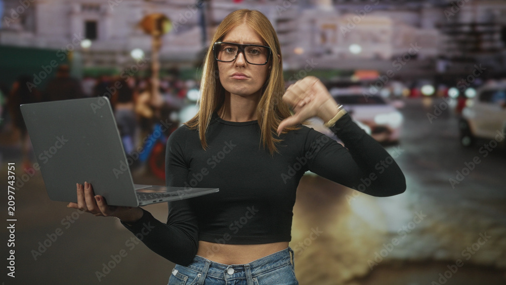 Fototapeta premium Woman holding laptop and pointing finger on a busy street in front of cars and buildings; frustration critique.