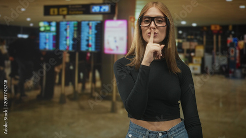 Slika na platnu Redhead woman wearing glasses presses index finger to lips for silence in a busy airport terminal check in area; playful secrecy