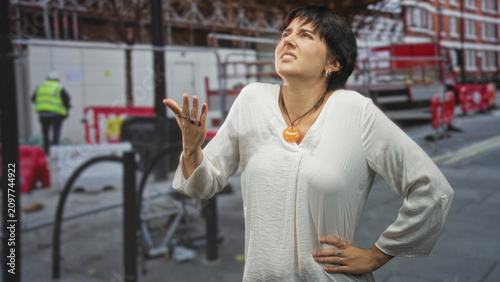 Quadro su tela Woman in white blouse with orange pendant, hand on hip and raised hand looking up on street; annoyance waiting
