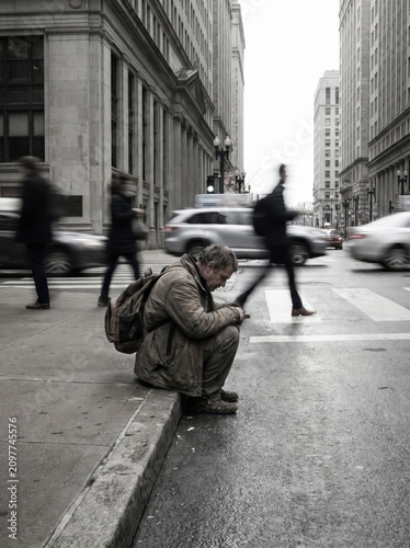 homeless man sitting on city street curb while blurred pedestrians rush past. urban isolation, poverty and inequality concept. desaturated dramatic scene. social issue, charity poster.