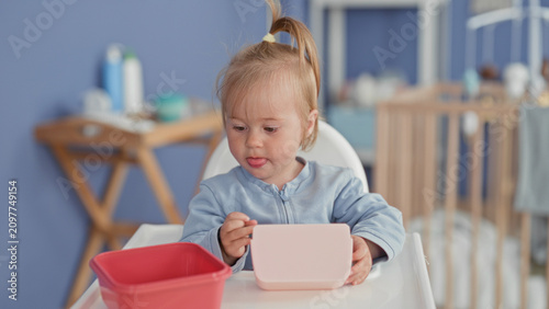 Foto Toddler girl eating with spoon at highchair, holding spoon with hand, pink container on tray and crib in view in building; concentration curiosity