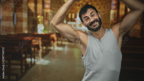 Schilderij op canvas Middle aged hispanic bearded man raises arms overhead showing biceps and armpit in a church aisle with pews and stained glass; joyful worship
