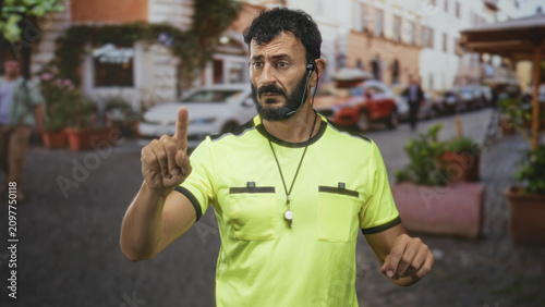 Foto Man referee in neon yellow shirt pointing index finger with whistle on necklace and earpiece, beard visible in street; authority control focus