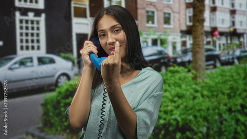 Photography Woman holding blue handset with middle finger raised while talking on corded phone on street near parked car and hedge; sassy defiance