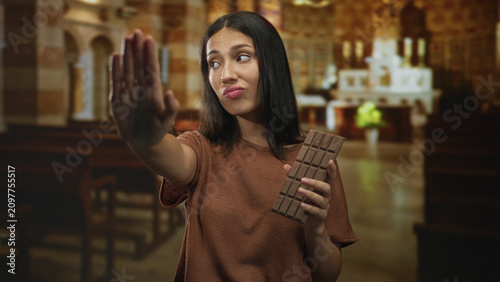 Bild auf Leinwand Woman holding chocolate bar, palm out stop gesture toward camera in church building with pursed lips and closed eyes; temptation restraint