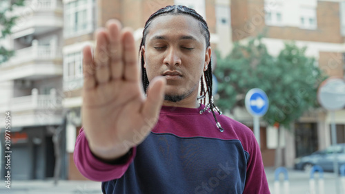 Obraz na plátně Man showing outstretched palm stop gesture on urban street with braided hair and sweatshirt; boundary defiance calm