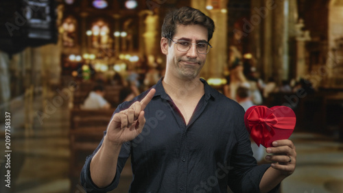 Принти на полотні Man holds red heart gift and points index finger in ornate building, showing hesitant thumb gesture; regret doubt
