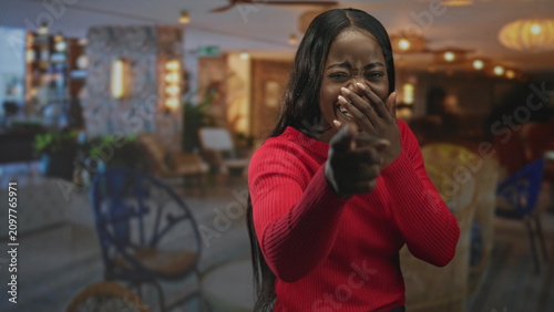 Photography Woman points finger like a finger gun and covers mouth with hand in a cafe lounge, red sweater visible; playful teasing