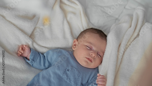 Canvastavla Boy newborn sleeping with arms raised and open hands on bed atop soft white blanket, wearing blue knitted onesie with elephant embroidery; serenity