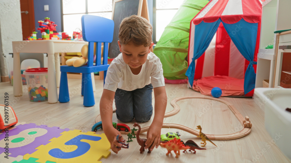 Fototapeta premium Boy kneeling and reaching with hand toward toy train on wooden floor in building playroom with tent, chair and foam puzzle mat; imaginative play joy.