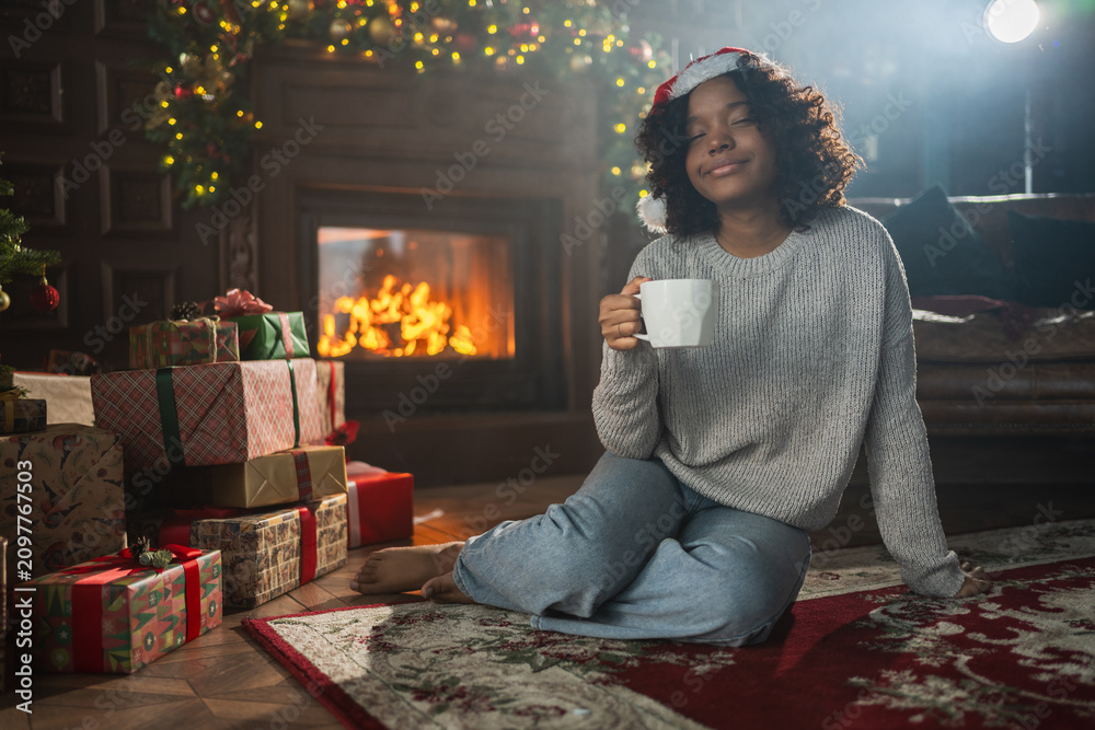 Obraz premium Merry Christmas. African American woman in Santa hat with cup of hot drink coffee tea near Christmas tree in dark interior. Girl in living room with Christmas tree and fireplace. Christmas eve at home