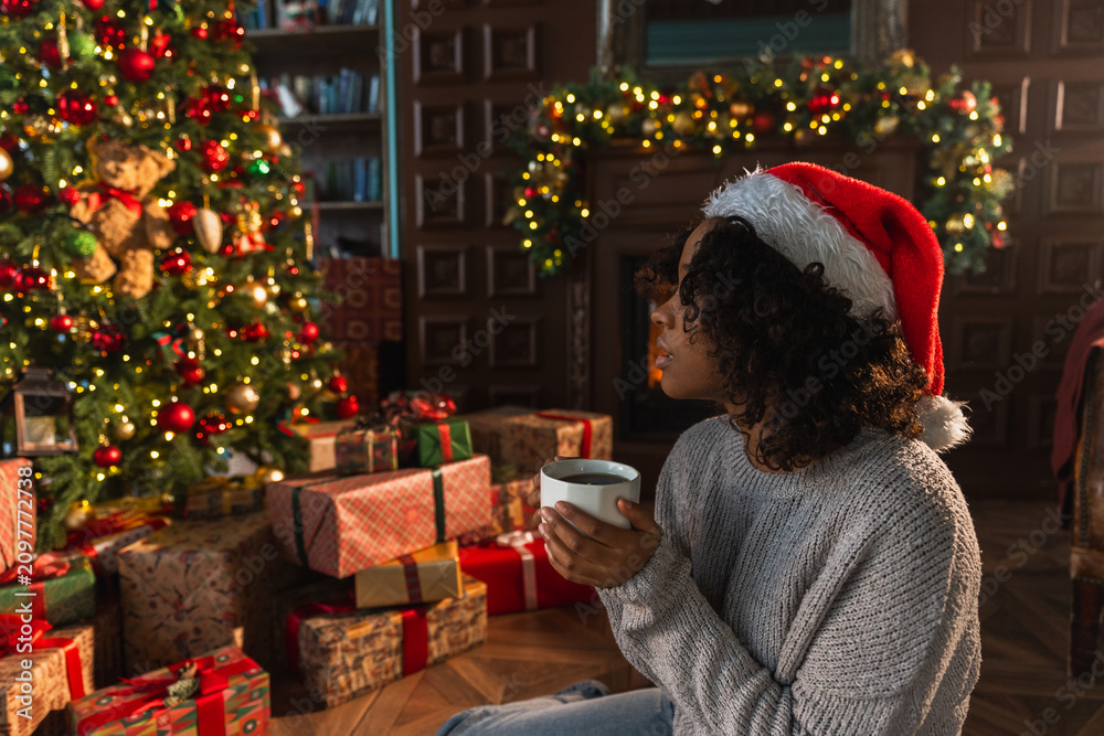 Obraz premium Merry Christmas. African American woman in Santa hat with cup of hot drink coffee tea near Christmas tree in dark interior. Girl in living room with Christmas tree and fireplace. Christmas eve at home