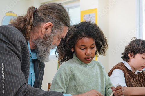 Quadro su tela Baby boomer teacher leans in to mentor two Generation Alpha pupils during handwr