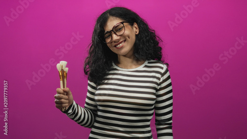 Принти на полотні Young arab brunette woman wearing glasses holding makeup brushes in one hand against a pink backdrop in studio, smiling and posing; playful beauty creativity