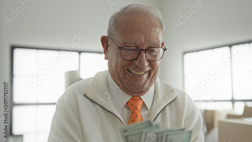 Fotografie Man counting dollars with hands and smiling expression amid stacked boxes and windows in a building interior, wearing glasses and an orange tie; financial joy