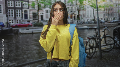 фотографія Teenage girl in yellow sweater with blue backpack covers mouth with hand on street by a canal and bikes; nervousness uncertainty