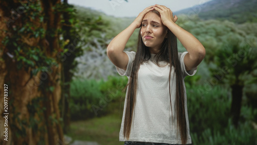 Canvastavla Young woman with hands on head crying in forest, wearing a white tshirt and long hair, standing among trees; sadness isolation vulnerability