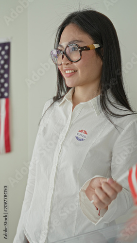 Foto Woman holds small american flag and displays a voted sticker by a transparent ballot box in a polling building; pride civic duty