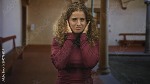 Fotografia Young woman pressing hands to ears with eyes closed, curly hair, wearing a maroon sweater, near benches and stone columns in a building; discomfort