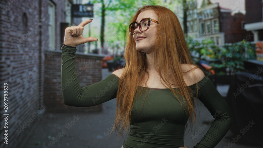 Fototapeta premium Redhead woman pinches fingers to indicate small size with her hand on a street, smiling broadly in glasses and green shirt; playful confidence.