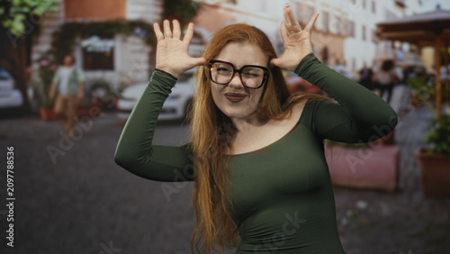 Foto Redhead woman in green top with glasses sticks out tongue and frames her face with both hands on a cobblestone street in front of shops; playful mischief