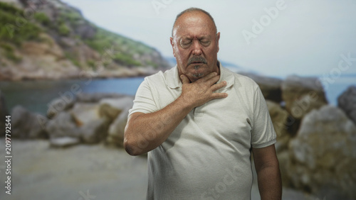 Photos Man holding throat with hand in choking gesture on rocky beach, eyes closed and head tilted back; distress