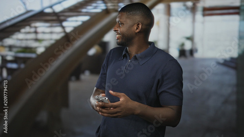Quadro su tela Man wearing navy polo shirt holding smartphone in hands, smiling and glancing sideways in airport terminal near staircase and concourse; contentment