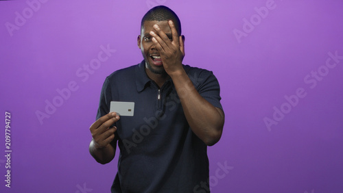 Foto Man holding creditcard and covering one eye with hand in a purple studio backdrop, mouth open in a startled expression; financial anxiety